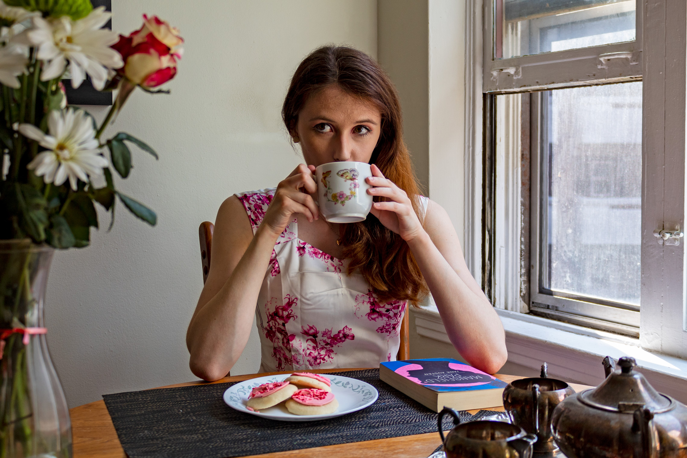 Woman sipping on a cup of tea; in front of her are cookies and a book