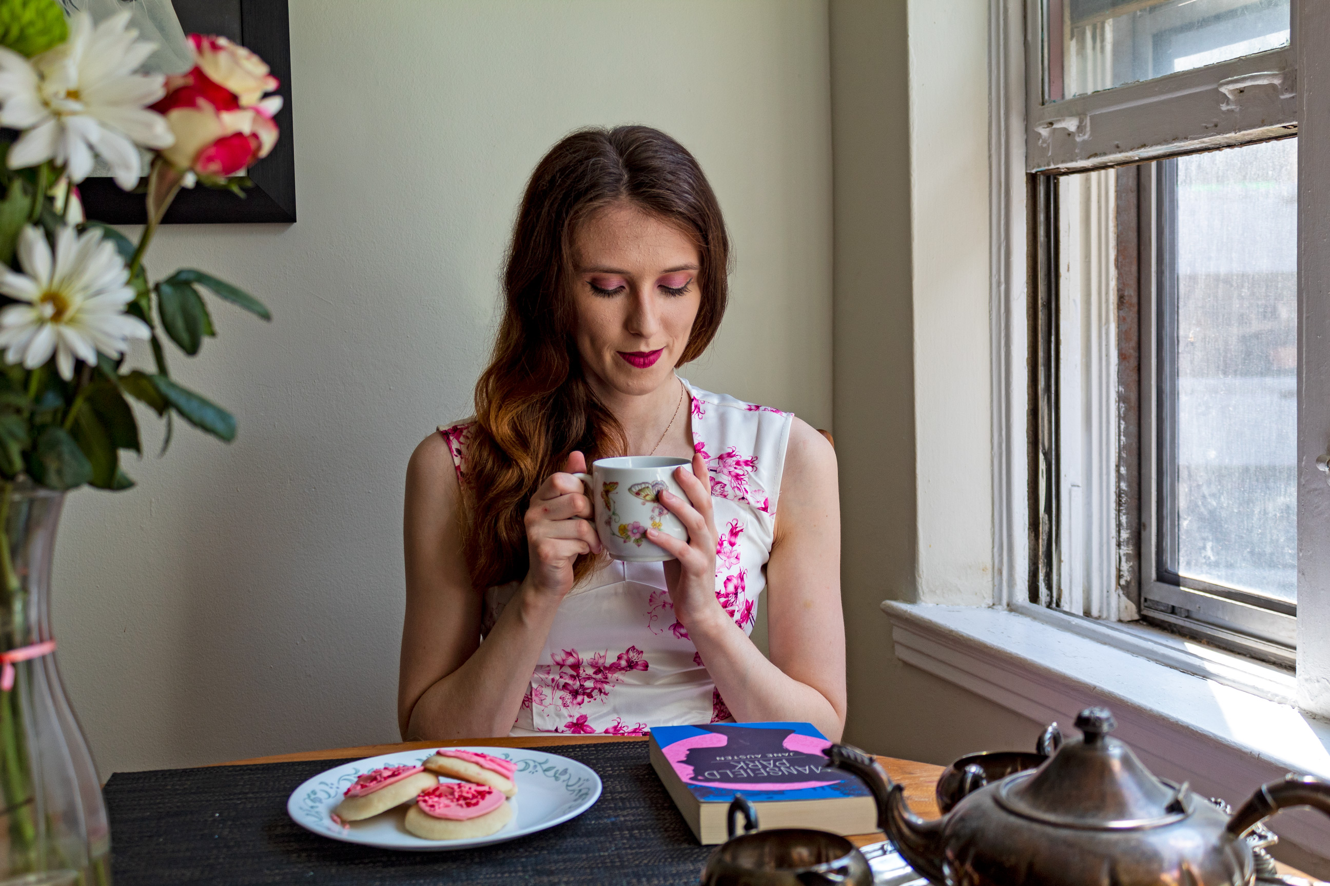 Woman looking down into the tea cup in her hand; in front of her are cookies and a book