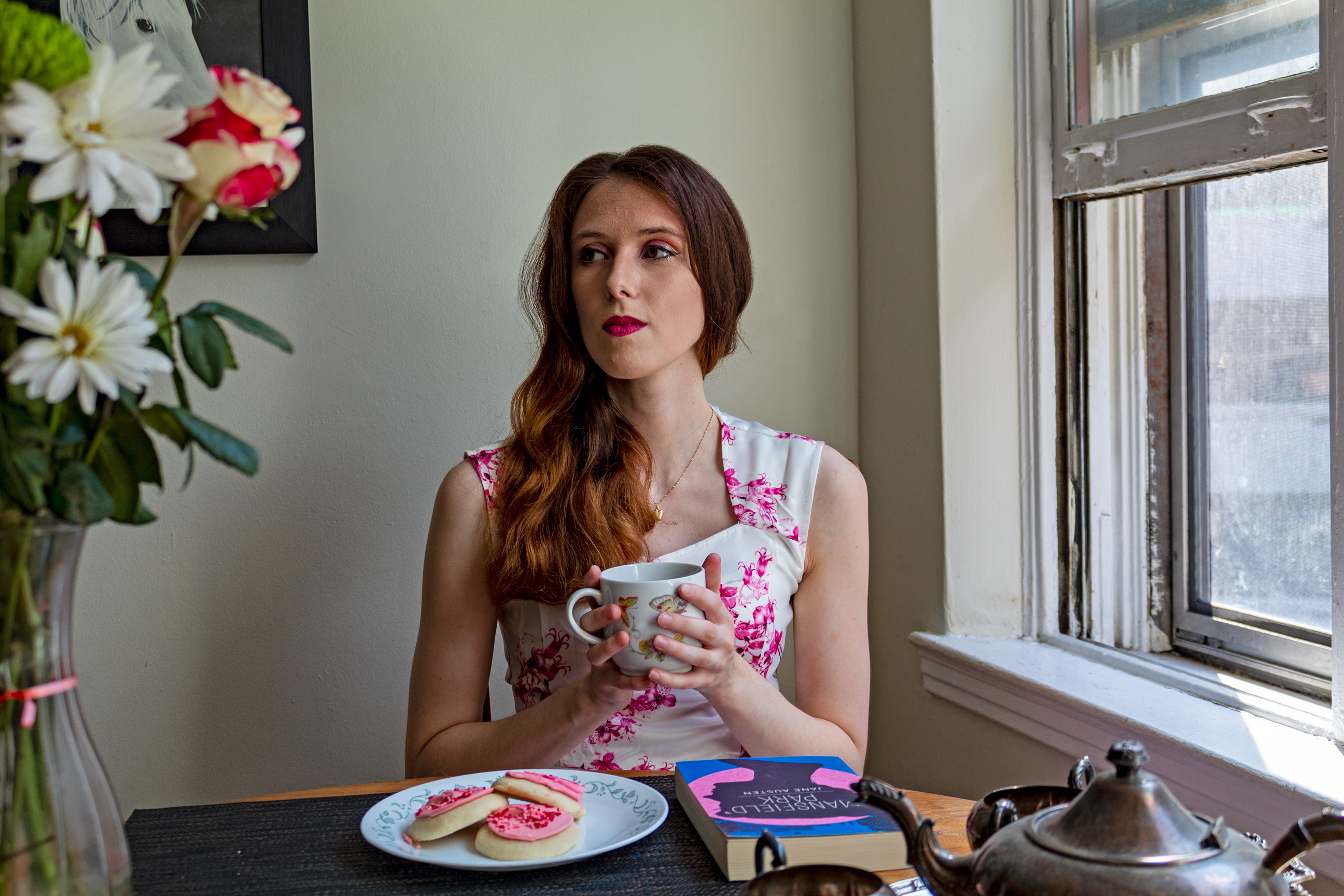 Woman holding a tea cup and gazing at a vase of flowers; in front of her are cookies and a book