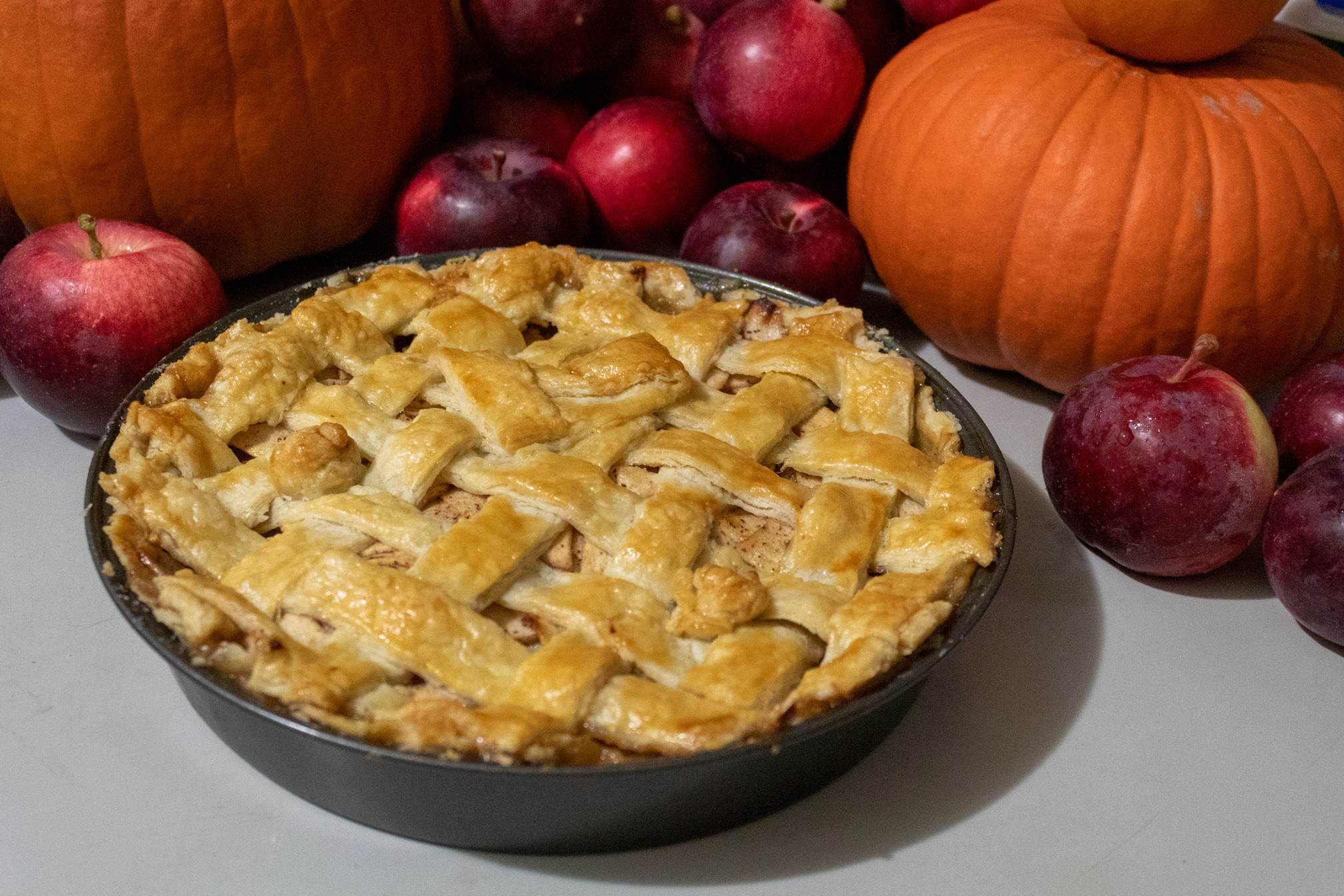 Apple pie with latticed top crust viewed from a skewed angle against a backdrop of Spartan apples and pumpkins